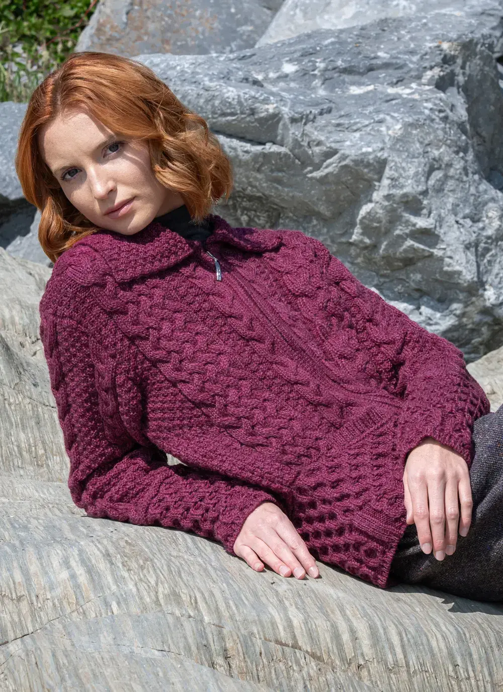 Red head model laying on rock in grace full zip aran cardigan in raspberry melange
